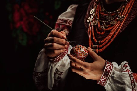Ukrainian woman painting traditional ornamets on Easter egg - pysanka. Artist working in national costume. Preparation for Christian holiday. 