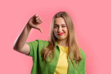 Portrait of unhappy european woman condemns with sign of dislike. Young millennial lady expressing discontent with showing thumbs-down gesture on pink studio background.