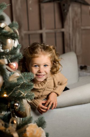 Photo for A beautiful laughing girl in a beige sweater sits in a light gray armchair and looks out playfully from behind a Christmas tree in a dark room - Royalty Free Image