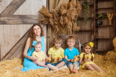 Téléchargez les photos : Image dépeint une famille d'une mère et de quatre enfants assis sur du foin dans une grange. Les enfants tiennent des canetons et portent des vêtements d'été jaunes et bleus. La mère a les cheveux ondulés. - en image libre de droit