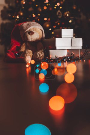 Photo for Cute Bear Dressed in Red Festive Santa Hat with Silver Gift Boxes Under Beautiful Glowing Christmas Tree. Happy New Year. - Royalty Free Image