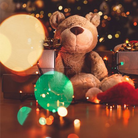 Foto de Bonito juguete de oso suave sentado cerca del hermoso árbol de Navidad festivo. Decorado con guirnalda brillante colorido. Acogedoras vacaciones de invierno en casa. - Imagen libre de derechos