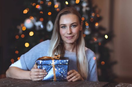 Téléchargez les photos : Portrait d'une jolie femme avec coffret cadeau festif à la maison sur fond d'arbre de Noël décoré. Joyeuses vacances d'hiver. - en image libre de droit