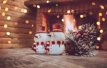 Foto de Juguetes festivos en casa de invierno decorada con festón. Dos muñeco de nieve y pino rama cerca de la chimenea. Nochebuena pacífica en casa. - Imagen libre de derechos