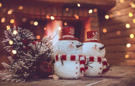 Foto de Juguetes festivos en acogedora cabaña de invierno. Dos muñeco de nieve y pino rama cerca de la chimenea. Nochebuena pacífica en casa. - Imagen libre de derechos