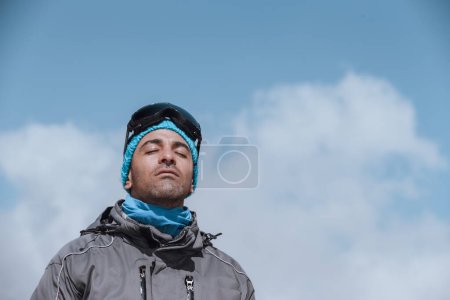 Téléchargez les photos : Portrait d'un bel homme aux yeux fermés de plaisir profitant d'une journée ensoleillée dans les montagnes sur fond de ciel bleu. Joyeuses vacances d'hiver. - en image libre de droit