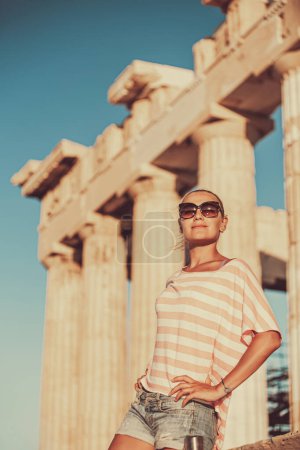 Foto de Retrato de una hermosa joven divirtiéndose en la soleada Grecia. Visitando ruinas antiguas magníficas en una Acrópolis ateniense, templo principal en Atenas antigua. - Imagen libre de derechos