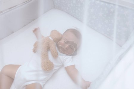 Foto de enfoque selectivo de un niño pequeño y lindo dormido en la cuna con un oso de juguete suave bajo un toldo suave blanco. Tiempo pacífico en casa.