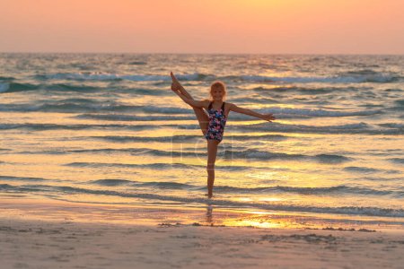 Foto de Linda chica adolescente haciendo gimnasia en la playa sobre el fondo del cielo puesta del sol. Vacaciones deportivas activas de verano. Infancia saludable. - Imagen libre de derechos