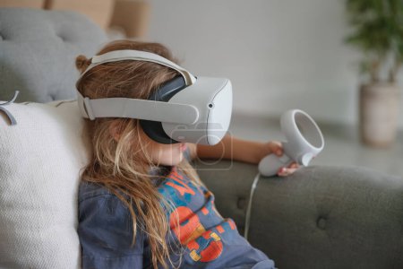 Téléchargez les photos : Photo d'un petit garçon jouant à un jeu vidéo à la maison. Technologie moderne. Simulation d'une réalité virtuelle. Un gamin intelligent qui aime le cyberespace. Masque VR - en image libre de droit