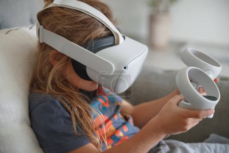 Téléchargez les photos : Photo d'un petit garçon portant un masque VR jouant à un jeu vidéo à la maison. Technologie moderne. Simulation d'une réalité virtuelle. Enfant intelligent profitant du cyberespace. - en image libre de droit