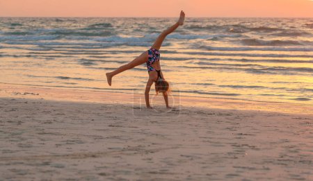 Photo for Cute teen girl doing gymnastics on the beach over sunset sky background. Active sportive summer holidays. Healthy childhood. - Royalty Free Image