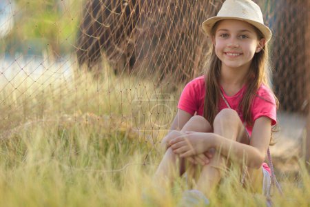 Téléchargez les photos : Jolie petite fille passant des vacances d'été à la campagne. Assis sur le terrain près du filet des pêcheurs. Profiter du temps à l'extérieur. - en image libre de droit