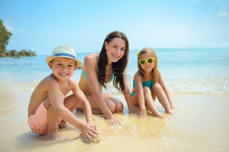 Young mother with two happy kids making a sand castle on the beach. Happy family spending summer vacation on the beach resort. People traveling.