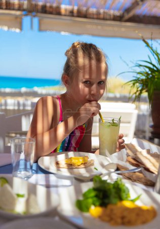 Photo for Portrait of a nice kid girl with pleasure drinking refreshing juice in the beach cafe. Enjoying summer holidays on resort. Happy tourist. - Royalty Free Image
