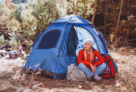 Foto de Mujer feliz con mochila sentada cerca de la tienda en el bosque montañoso. Vacaciones activas de campamento de otoño con amigos en las montañas. - Imagen libre de derechos