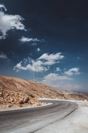 Téléchargez les photos : Photo d'une route sinueuse du désert, parmi les montagnes blanchies par le soleil sous un ciel bleu qui est dilué avec des nuages blanc neige. Road trip wanderlust. - en image libre de droit