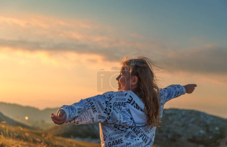 Foto de Feliz niña divirtiéndose en las montañas. Niño feliz con las manos levantadas disfrutando de la luz suave del atardecer. Campamento de verano en las montañas. - Imagen libre de derechos