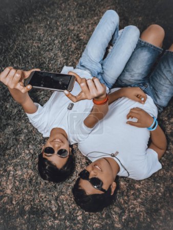 Foto de Dos buenos adolescentes con gafas de sol, sonriendo felizmente, tumbados en el suelo, tomando una foto memorable del momento, la amistad y las emociones. - Imagen libre de derechos