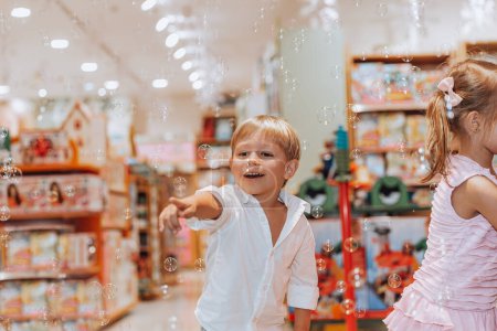 Foto de Bonito hermano pequeño y hermana con placer soplando burbujas de jabón en una tienda de juguetes. Niños alegres divirtiéndose en la habitación de los niños. Felicidad por todas partes. - Imagen libre de derechos