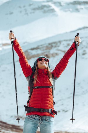 Photo for Pretty girl with raised up hands standing among beautiful snowy mountains with walking poles in hands. Looking in the sky. Enjoying winter holidays. - Royalty Free Image
