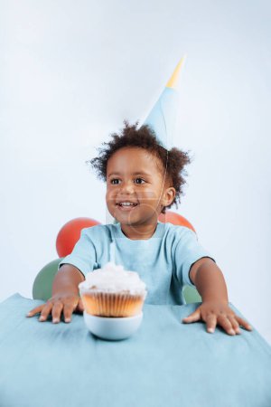 Foto de Un pequeño niño afroamericano alegre sonriente vestido con un sombrero festivo disfruta de su pastel de cumpleaños. Un símbolo de cumpleaños en todo el mundo. - Imagen libre de derechos
