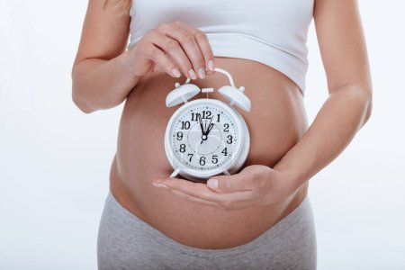 Téléchargez les photos : Une fille enceinte tient une montre ronde dans ses mains, comme si elle disait que très bientôt elle aura un bébé. Une partie du corps. Isolé sur fond blanc. - en image libre de droit
