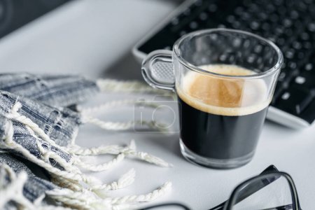 Téléchargez les photos : Une tasse de café chaud, fort et revigorant se trouve sur le bureau à côté de l'ordinateur et des lunettes. Matin classique pour les employés de bureau. Travail à distance. - en image libre de droit