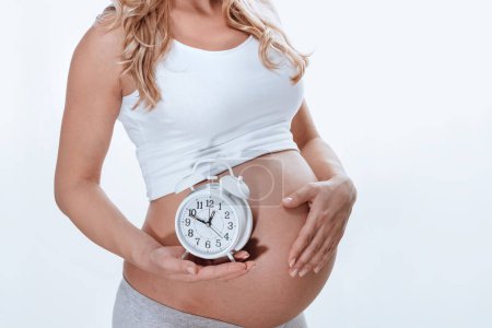 Téléchargez les photos : Une fille enceinte tient une alarme ronde comme si elle disait que très bientôt elle aura un bébé. Une partie du corps. Isolé sur fond blanc. - en image libre de droit