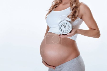 Photo for A pregnant girl stands sideways and holds a alarm clock in her hands. Very soon her little miracle will be born. New life concept. - Royalty Free Image