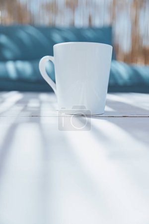 Téléchargez les photos : Une tasse blanche avec une boisson du matin se tient sur la table d'un bungalow d'été à côté d'un canapé bleu. Maison d'été confortable à la station. Concept d'ambiance estivale. - en image libre de droit