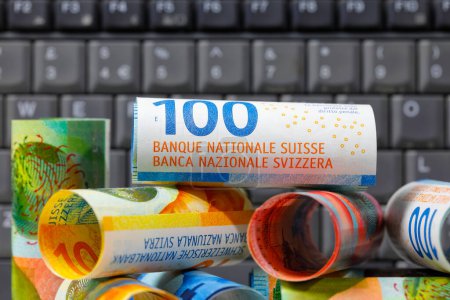 Foto de Billetes en franco suizo de varias denominaciones, apilados en una pila y colocados en el fondo de un teclado de ordenador. Papel moneda CHF, edición de billetes suizos, emitidos de abril de 2016 a 2019. - Imagen libre de derechos