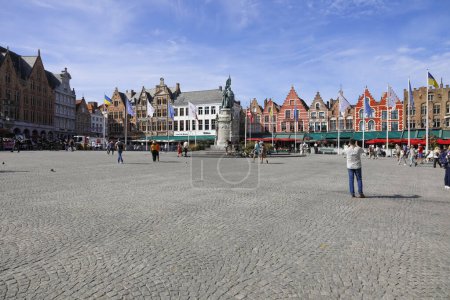 Foto de Brujas, Bélgica - 12 de septiembre de 2022: Las coloridas casas de ladrillo forman la fachada de la Plaza del Mercado. También hay una gran estatua de bronce casi en el centro de la plaza. - Imagen libre de derechos