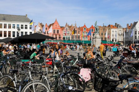Foto de Brujas, Bélgica - 12 de septiembre de 2022: Las coloridas casas de ladrillo forman la fachada de la Plaza del Mercado. Se puede ver gente en la plaza, estacionamiento para bicicletas, restaurantes al aire libre y una gran estatua de bronce - Imagen libre de derechos