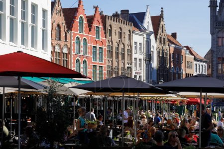 Foto de Brujas, Bélgica - 12 de septiembre de 2022: Las coloridas casas de ladrillo forman la fachada de la Plaza del Mercado. Se puede ver a la gente bajo grandes sombrillas repartidas en un restaurante al aire libre - Imagen libre de derechos