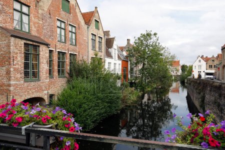 Foto de Brujas, Bélgica - 13 de septiembre de 2022: Hay casas de ladrillo junto al canal, hay mucha vegetación y en la barandilla del puente, hay flores en macetas - Imagen libre de derechos