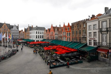 Foto de Brujas, Bélgica - 15 de septiembre de 2022: Casas coloridas seguidas en la fachada de Market Square con muchas sombrillas sobre restaurantes al aire libre adyacentes - Imagen libre de derechos