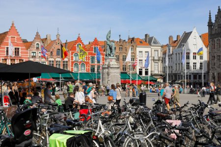 Foto de Brujas, Bélgica - 12 de septiembre de 2022: Las coloridas casas de ladrillo forman la fachada de la Plaza del Mercado. Se puede ver gente en la plaza, estacionamiento para bicicletas, restaurantes al aire libre y una gran estatua de bronce - Imagen libre de derechos