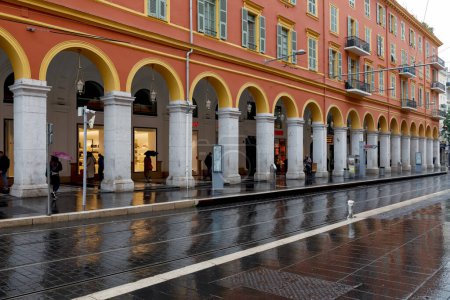 Téléchargez les photos : Nice, France - 30 avril 2023 : Le bâtiment avec l'arcade et le tramway local s'arrêtent à la place Massena par un jour de pluie. La chaussée et les voies de tramway sont mouillées par la pluie. - en image libre de droit