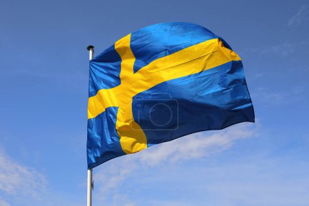 The national flag of Sweden, flying on a flagstaff against a blue sky on a sunny day