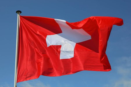 The national flag of Switzerland flying from a flagstaff against a blue sky on a sunny day.