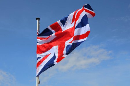 The flag of the United Kingdom waving on a flagstaff against a blue sky on a sunny day.
