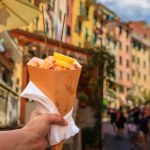 Hand holding a cone of fritto misto, Italian street food mix of fried calamari and shrimp with colorful houses in Riomaggiore, Cinque Terre Italy