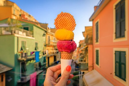 Woman s hand holding an artisanal gelato with a cookie and a view of traditional colorful houses in Manarola old town, Cinque Terre, Italy
