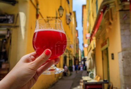 Téléchargez les photos : Main de femme tenant un verre de framboise Lambic ale dans un restaurant en plein air avec un fond de bâtiments flous dans la vieille ville Nice, Sud de la France - en image libre de droit