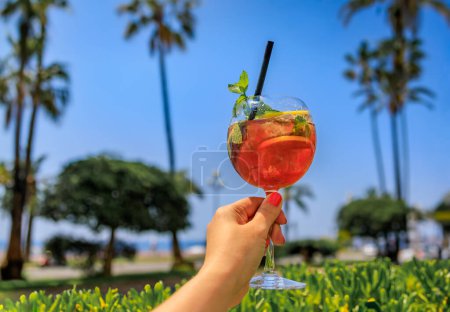 Téléchargez les photos : Main de femme tenant un cocktail de fantaisie dans un bar extérieur sur la Promenade des Anglais avec un fond de palmiers et de la mer Méditerranée à Nice, France - en image libre de droit