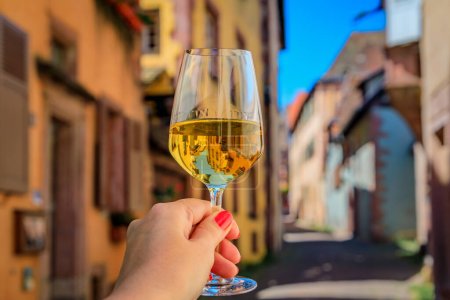 Foto de La mano de la mujer con una copa de vino blanco en un restaurante al aire libre, borrosa casas de entramado de madera en Riquewihr, Francia, un pueblo en la Ruta del Vino de Alsacia - Imagen libre de derechos