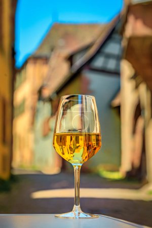 Foto de Una copa de vino blanco local en un restaurante al aire libre con casas borrosas de entramado de madera en Riquewihr, Francia, un pueblo en la Ruta del Vino de Alsacia - Imagen libre de derechos