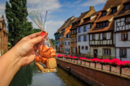 Foto de Mano de mujer sosteniendo una bolsa de galletas artesanales con casas de madera sobre los canales del río Lauch en Petite Venise, Colmar, Alsacia, Francia - Imagen libre de derechos