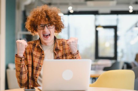 Foto de Buenas noticias. Adolescente masculino expresando éxito frente a la computadora portátil en la cafetería, apretando el puño y gritando, espacio para copiar - Imagen libre de derechos